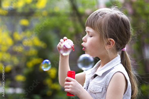 Profile of pretty small preschool blond girl with funny serious expression blowing colorful transparent soap bubbles outdoors on blurred green summer background. Joy of careless childhood concept.