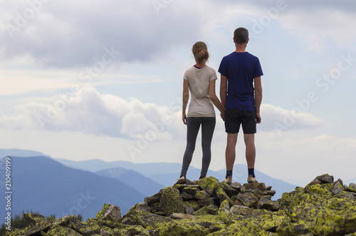 Back view of young tourist couple, athletic man and slim girl stand holding hand on rocky mountain top enjoying breathtaking summer mountain panorama. Tourism, traveling and healthy lifestyle concept.