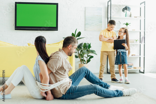 parents sitting tied with rope on floor and children looking at clipboard at home