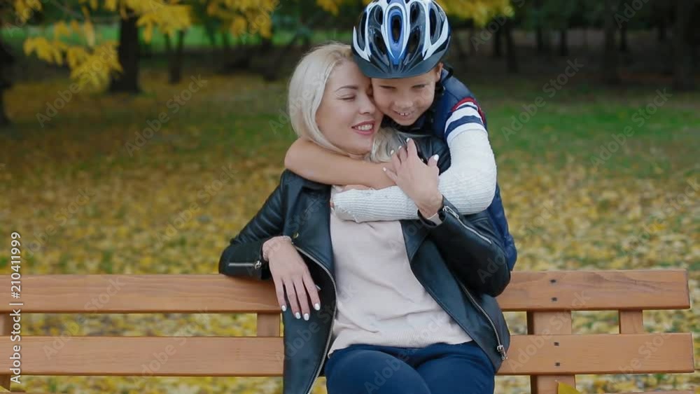 Young caucasian woman sitting on the bench. Suddenly her some comes to her from the back and cuddle her. Mother and son spending nice time together. The child wear helmet and his arm is broken.