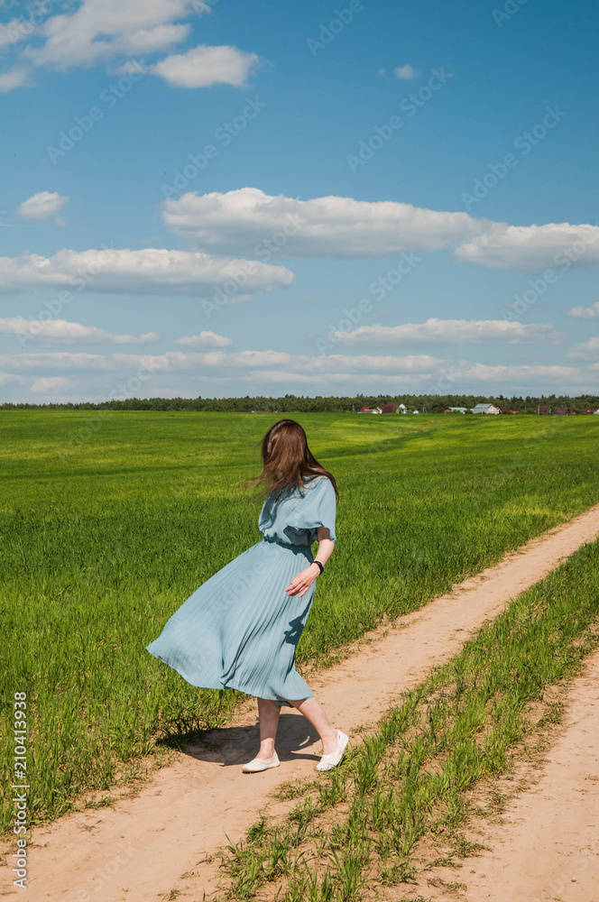 Beautiful young romantic girl outdoor in a blue dress with long hair enjoying nature, walking, spinning and dancing on the green field in the village