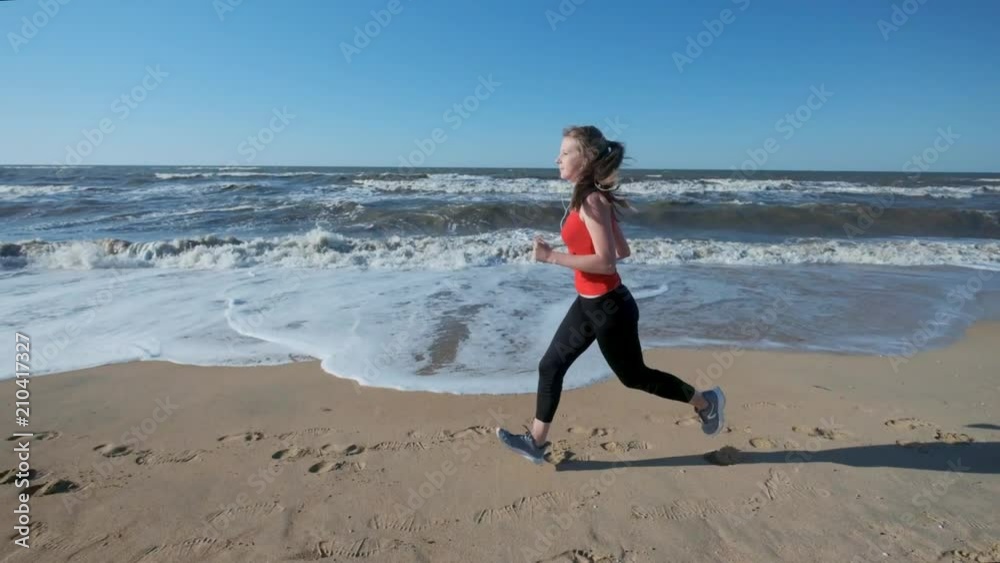 Portrait: a young girl runs along the beach in red sportswear. Listens to music on headphones. Performs an evening or morning jog at sunset or dawn Portrait: a young girl runs along the beach in red sportswear. Listens to music on headphones. Performs an evening or morning jog at sunset or dawn
