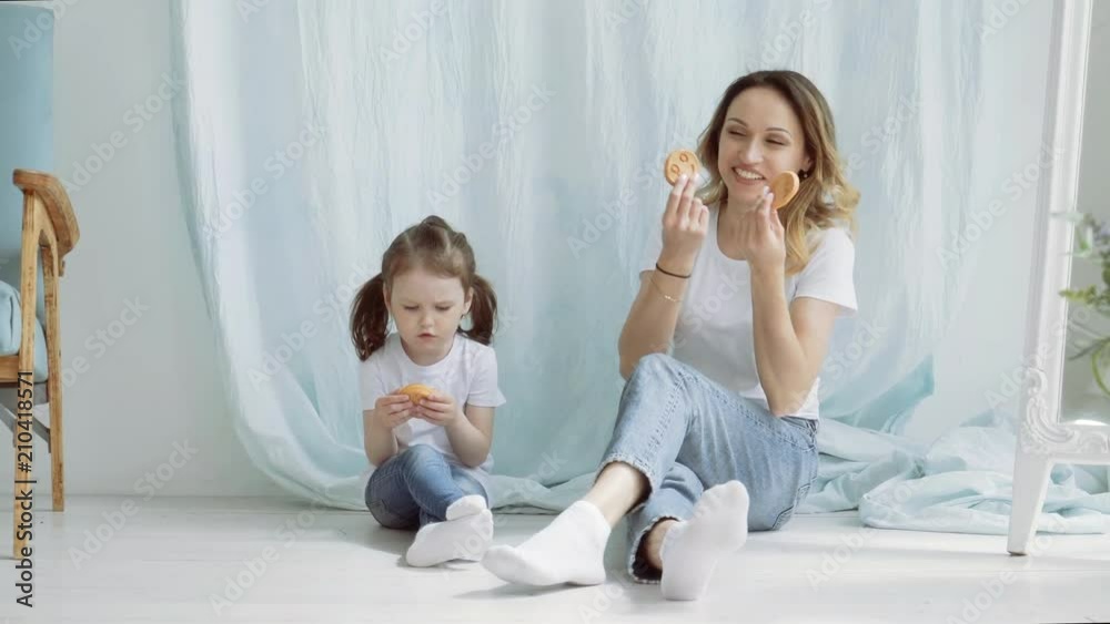 A pretty woman with a little girl with cookies in her hands is sitting on the floor and talking.