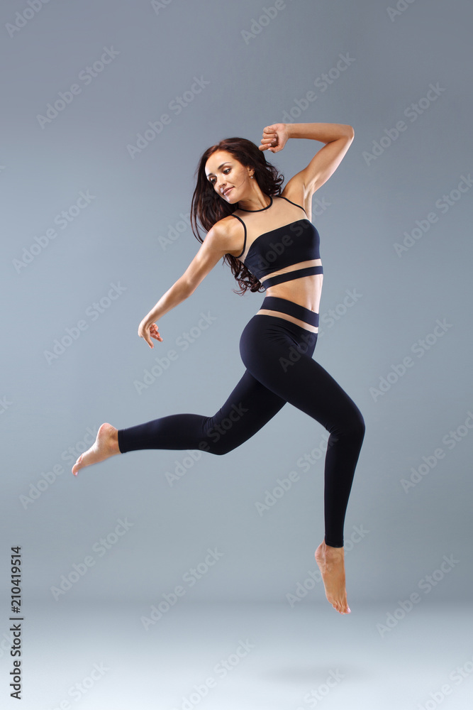 Attractive excited fitness girl, ballerina dancer in sportwear jumping of joy isolated over gray background.