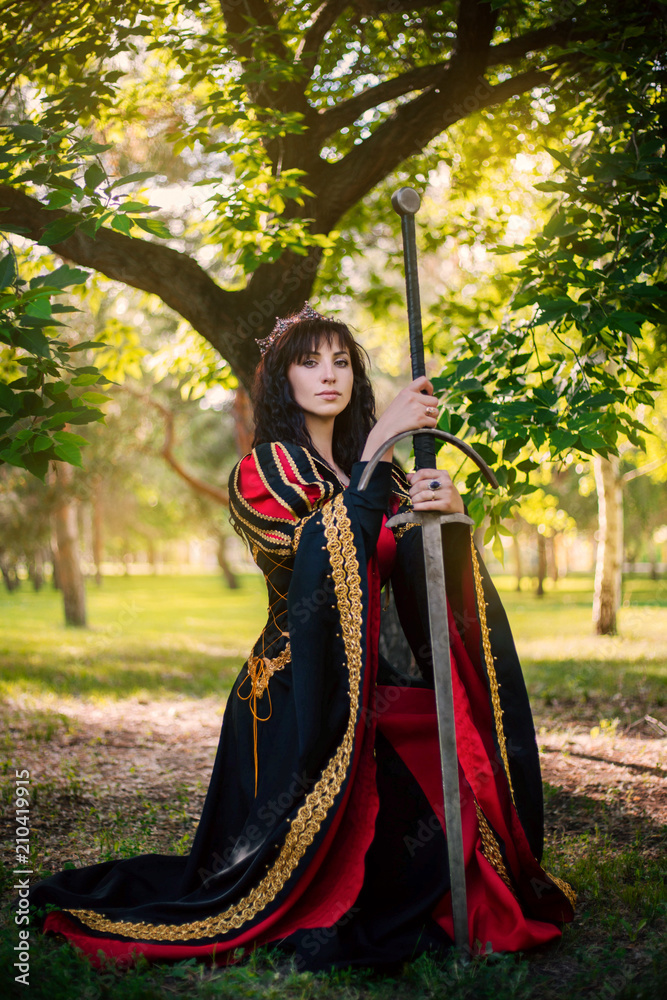 The Queen with a sword in her hands. Beautiful girl in the crown. Fantasy. Medieval black and red dress with gold embroidery. The Queen with a sword in her hands. Beautiful girl in the crown. Fantasy. Medieval black and red dress with gold embroidery.