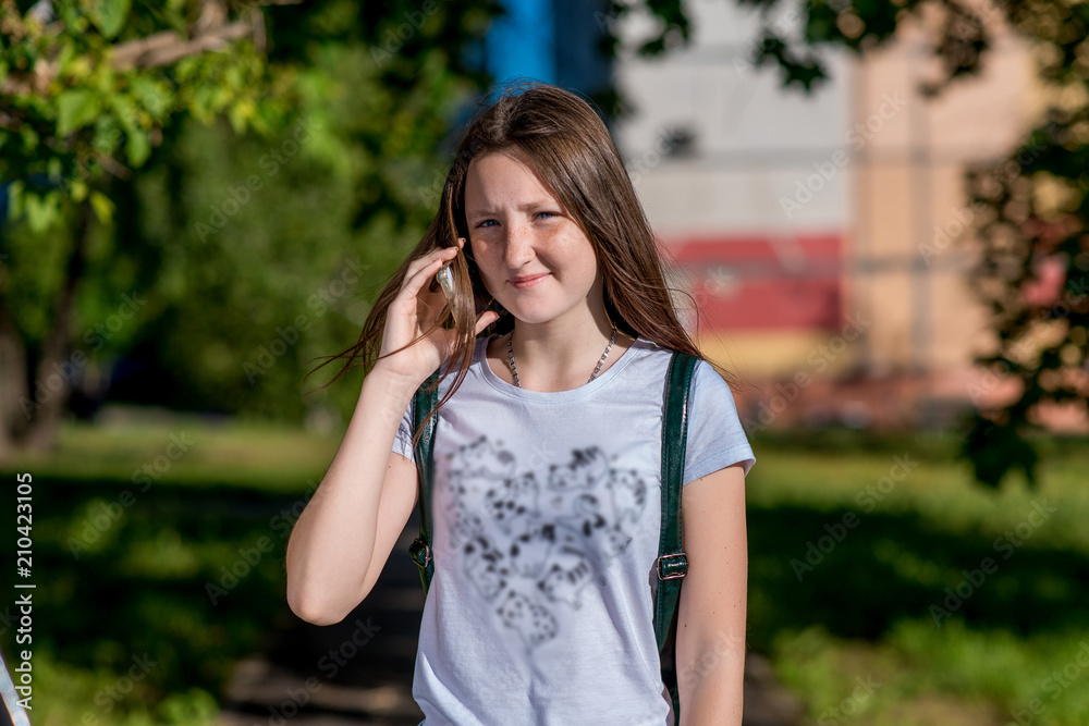 Girl schoolgirl. Summer in nature. Holds the hands of a smartphone. She calls on phone. Smiles happily. In the summer In the bright sun. In a white T-shirt behind his backpack. Girl schoolgirl. Summer in nature. Holds the hands of a smartphone. She calls on phone. Smiles happily. In the summer In the bright sun. In a white T-shirt behind his backpack.