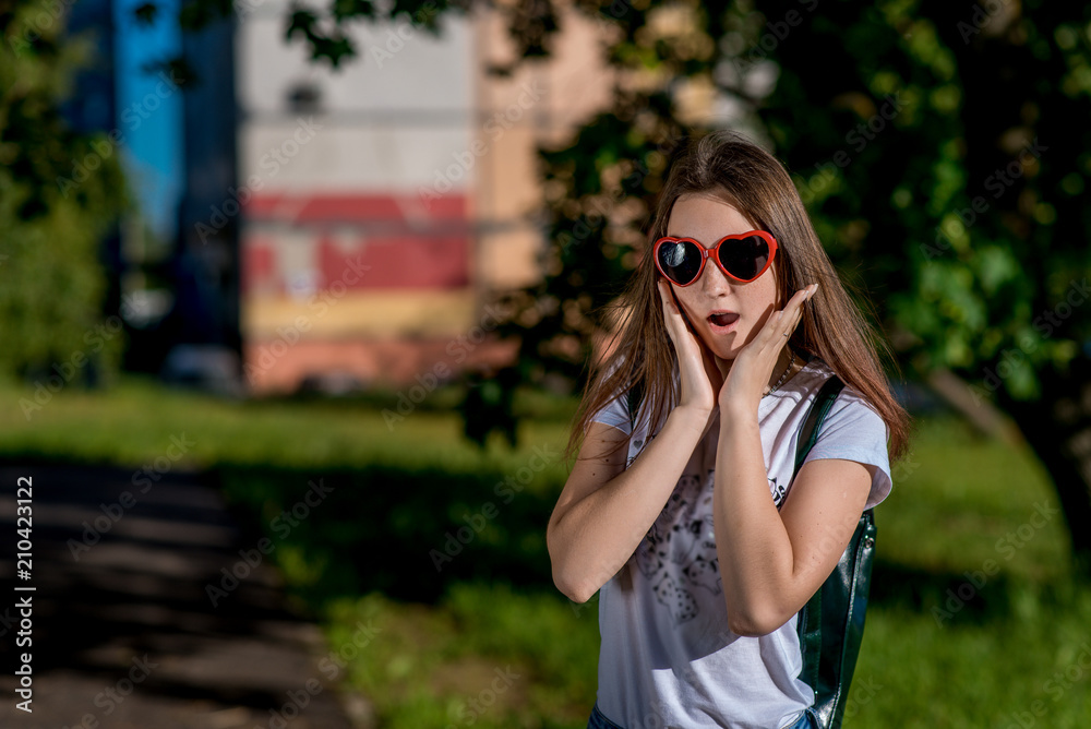 Girl schoolgirl in summer in city. In the glasses in the shape of the heart behind the backpack. Happy smiling. The concept of surprise surprise surprise. Free space for text. Girl schoolgirl in summer in city. In the glasses in the shape of the heart behind the backpack. Happy smiling. The concept of surprise surprise surprise. Free space for text.