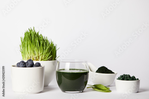 fresh smoothie from spirulina, cups with spirulina grass and blueberries, leaves, bowls with spirulina powder and spirulina pills on grey background