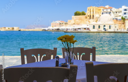 Empty greek cafe, white tablecloth, blue furniture and flowers on the table, depth of field blur background on Crete Island, Greece