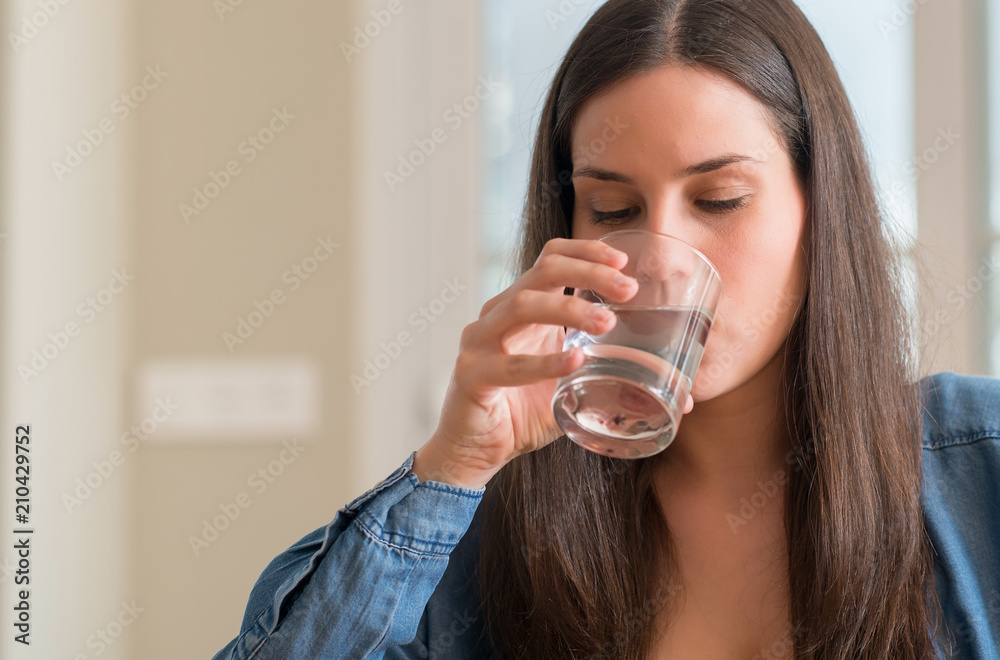 Young woman drinking glass of water at home with a confident expression on smart face thinking serious Young woman drinking glass of water at home with a confident expression on smart face thinking serious