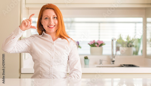 Redhead woman at kitchen smiling and confident gesturing with hand doing size sign with fingers while looking and the camera. Measure concept.