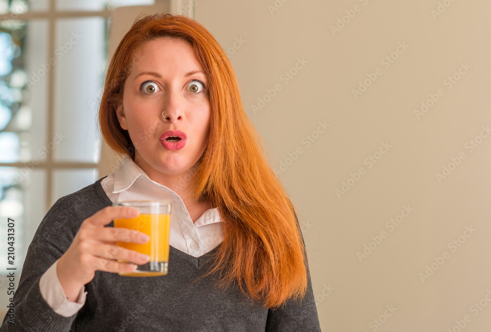 Redhead woman holding orange juice glass at home, scared in shock with a surprise face, afraid and excited with fear expression Redhead woman holding orange juice glass at home, scared in shock with a surprise face, afraid and excited with fear expression