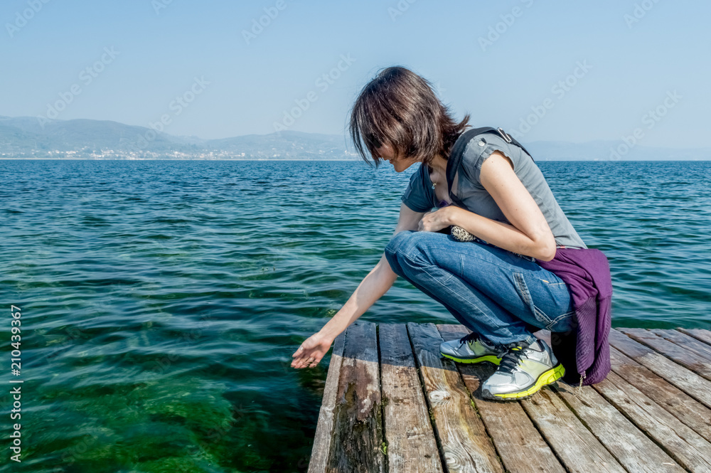 Beautiful young woman happily splashing the lake water with her hand on a wooden pier