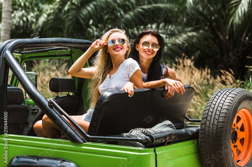 two girls best friends travel on a tropical island in a car with an open top are having fun having a smile. enjoyment travel, excursion summer dressed shorts T-shirts sunglasses