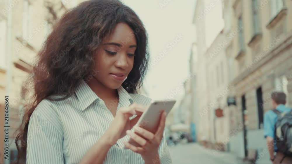 Smiling attractive Afro-American woman taps screen of her phone, typing sms, sending message. Browsing the internet, social networks, blogger, positive emotions. Rotation view Smiling attractive Afro-American woman taps screen of her phone, typing sms, sending message. Browsing the internet, social networks, blogger, positive emotions. Rotation view