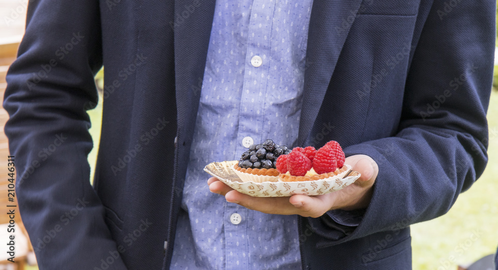 A man in a suit holds a paper bowl of sweet fruit desserts..Pastry - sweet cups with raspberries and the other one with blueberries..Food shopping during garden party, farmers food market.