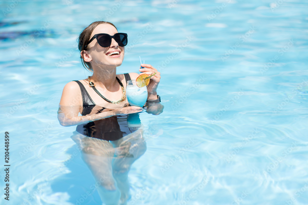 Beautiful woman in black swimsuit and sunglasses swimming with cocktail in the water pool outdoors