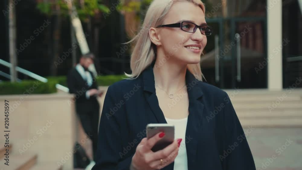 Smiling beautiful young woman with glasses walks down the central city’s street uses phone portrait business fashion hand cellphone internet messaging smartphone text happy reading slow motion Smiling beautiful young woman with glasses walks down the central city’s street uses phone portrait business fashion hand cellphone internet messaging smartphone text happy reading slow motion