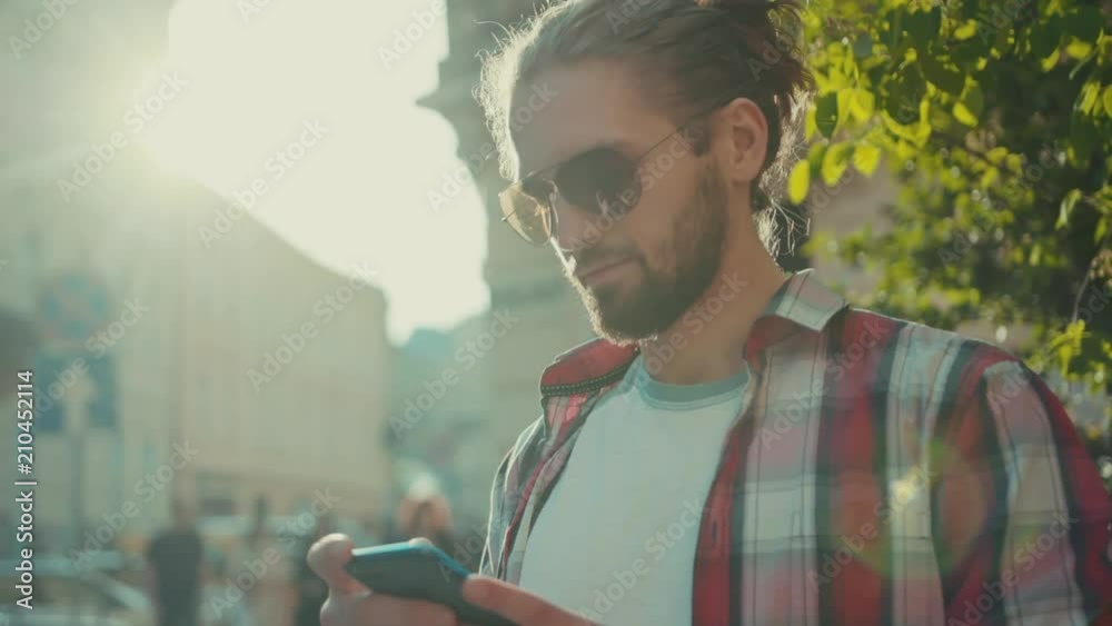 Young stylish young man with sunglasses standing using phone street texting casual clothing shirt city looking around sunny sunlight elegant trendy perfect look portrait slow motion close up