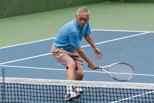 Healthy Chinese elderly man showing flexibility while reach for the low tennis backhand volley.