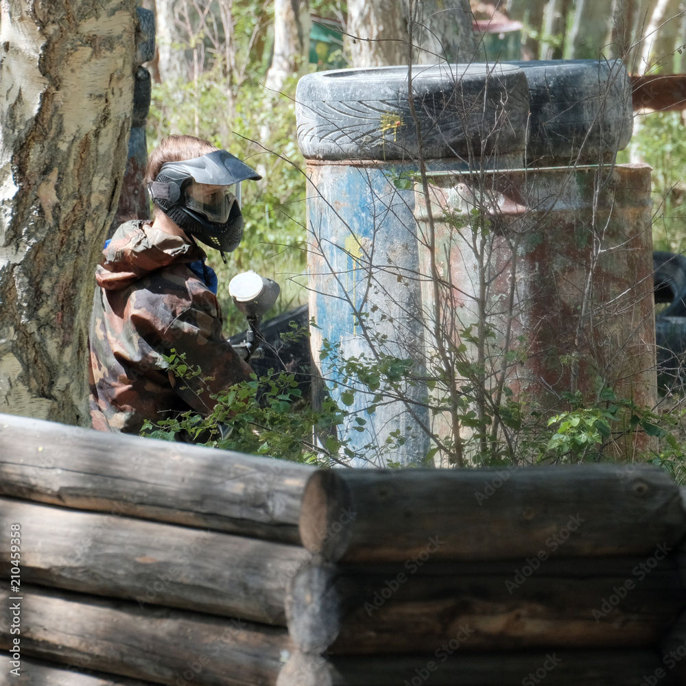 Obraz premium Extreme team sport. The player in the paintball looks out of hiding. Camouflage suit, protective mask and weapons. Tires, barrels, wooden fences in the forest.
