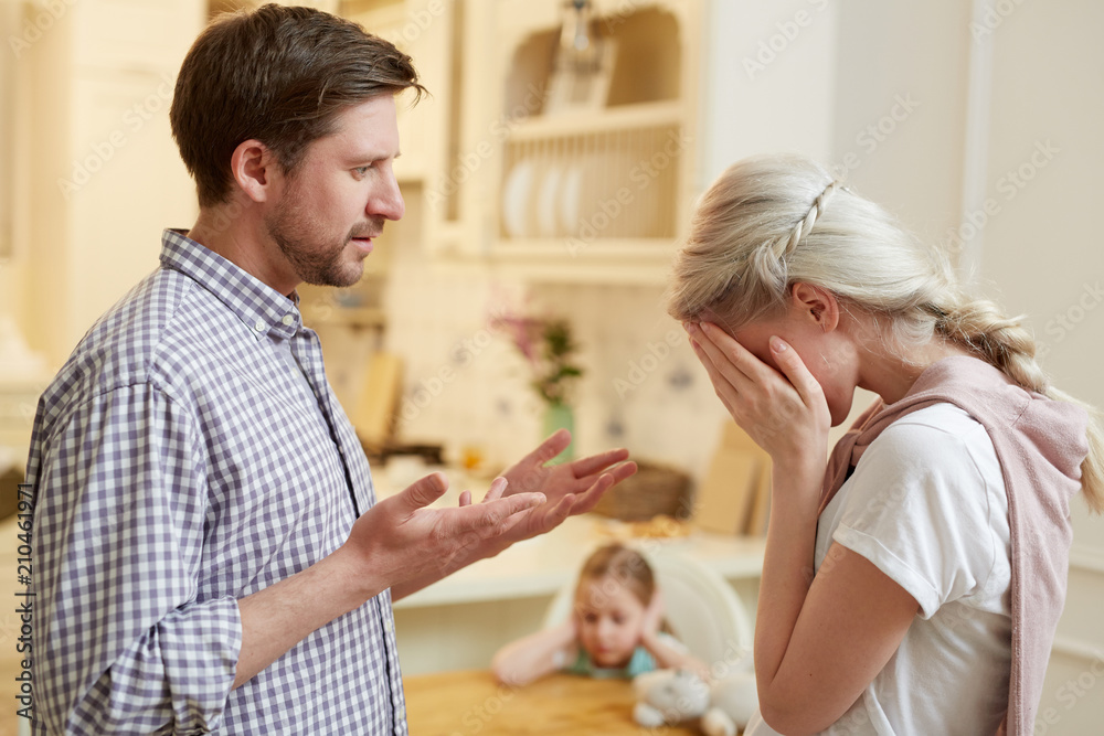 Annoyed husband expressing his displeasure to crying wife with their little daughter on background Annoyed husband expressing his displeasure to crying wife with their little daughter on background