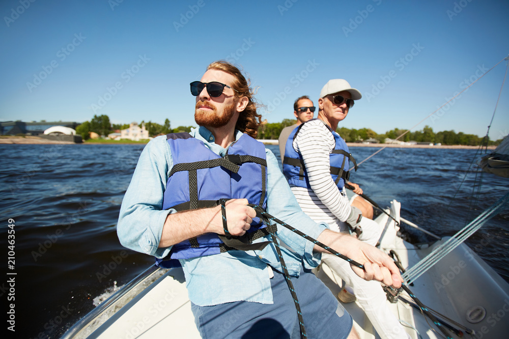 Fototapeta premium Group of men in protective lifejackets and sunglasses floating on yacht or motor boat on summer day