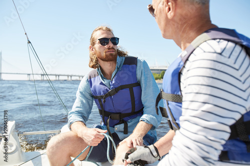 Young and senior active men in lifejackets sailing on yacht and talking on hot summer day