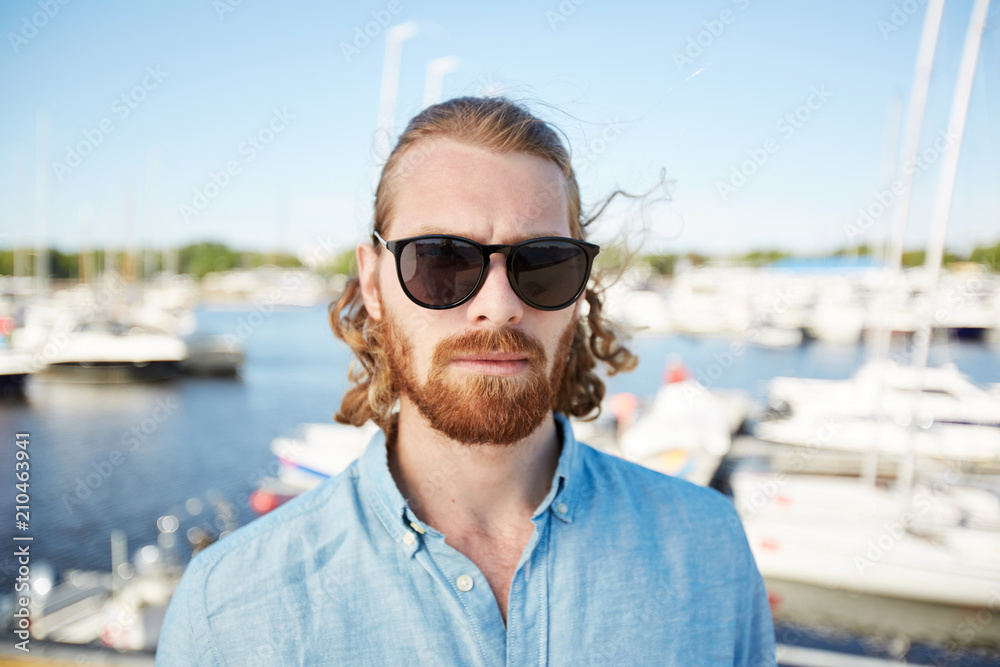Bearded man in sunglasses and denim shirt standing in front of camera with yachts on background Bearded man in sunglasses and denim shirt standing in front of camera with yachts on background