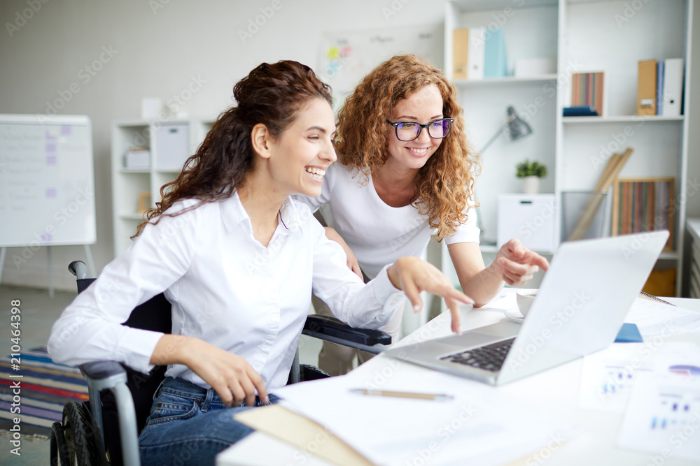 Happy woman in wheelchair pointing at laptop display while explaining online data to colleague at meeting Happy woman in wheelchair pointing at laptop display while explaining online data to colleague at meeting