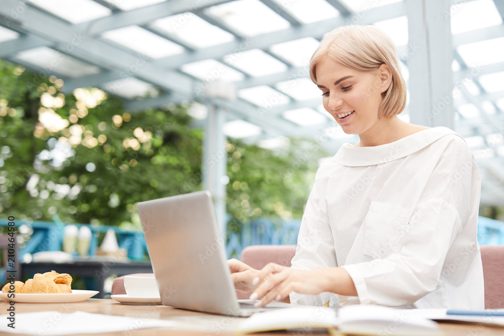 Happy blond businesswoman sitting in front of laptop and typing on keypad in oranjery Happy blond businesswoman sitting in front of laptop and typing on keypad in oranjery