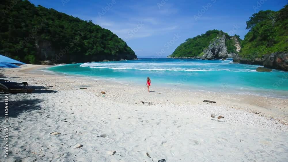 Young beautiful woman in red swimsuit staying and watching on exotic beach surrounded by green hills, Nusa Penida island, Indonesia Young beautiful woman in red swimsuit staying and watching on exotic beach surrounded by green hills, Nusa Penida island, Indonesia