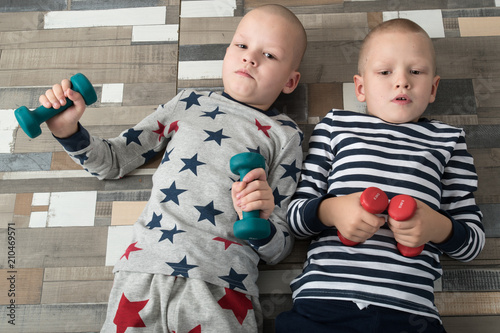 Two funny boys in sports clothes raise dumbbells up. Children are actively engaged in fitness at home. Copy space photo