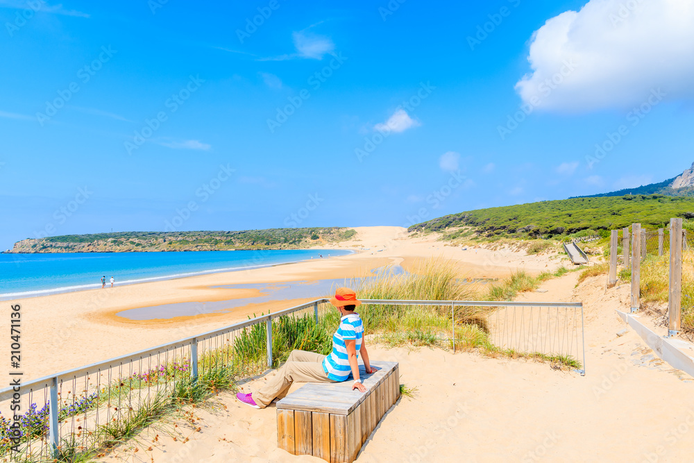 Naklejka premium Young woman tourist sitting on bench and looking at idyllic white sand Bolonia beach, Andalusia, Spain