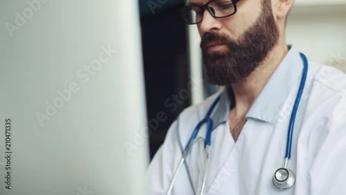 Brown-haired Caucasian physician in spectacles sitting at table and looking at monitor. Content man with beard typing on keyboard of computer at clinic. Indoors.
