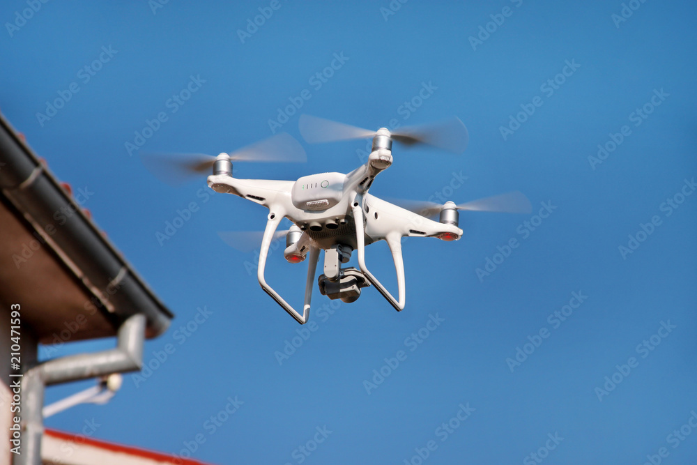 Drone quad copter fly on blue sky in background. Modern drone is flying in air, to take photos and record footage from above. Drone with four propellers, camera and red warning lights. New technology. Drone quad copter fly on blue sky in background. Modern drone is flying in air, to take photos and record footage from above. Drone with four propellers, camera and red warning lights. New technology.