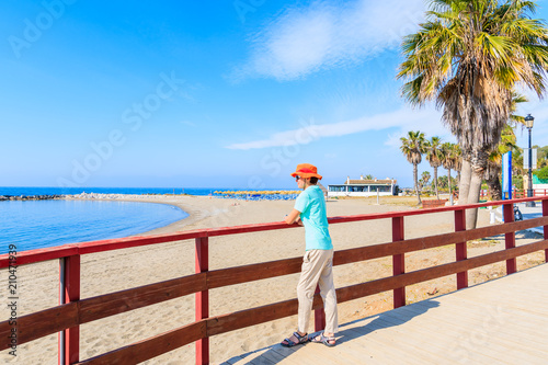 Young woman tourist standing on coastal promenade and looking at beautiful beach with palm trees near Marbella, Andalusia, Spain