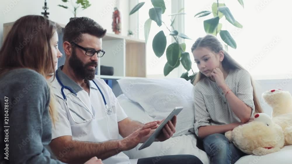 Caucasian doctor in spectacles touching digital tablet and talking to girls. Mother and ill daughter looking at screen. Communication. Indoors. Caucasian doctor in spectacles touching digital tablet and talking to girls. Mother and ill daughter looking at screen. Communication. Indoors.