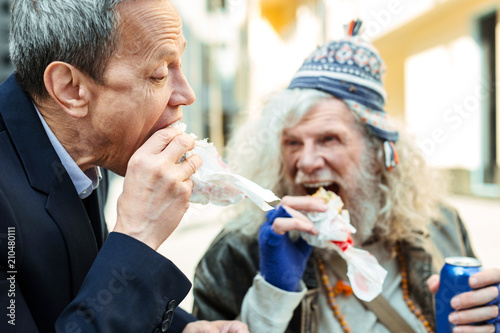 Nice refugee. Caring businessman spending his break time with nice kind-hearted starving refugee while eating burger together