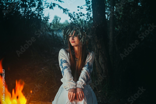 Woman in ethnic clothes with chaplet on a head sitting near bonfire. Concept of Pagan holiday. photo