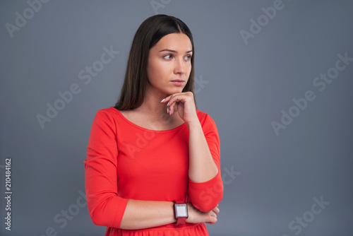 Weighing pros and cons. Charming young woman in a red dress thinking about something and resting her chin on her hand