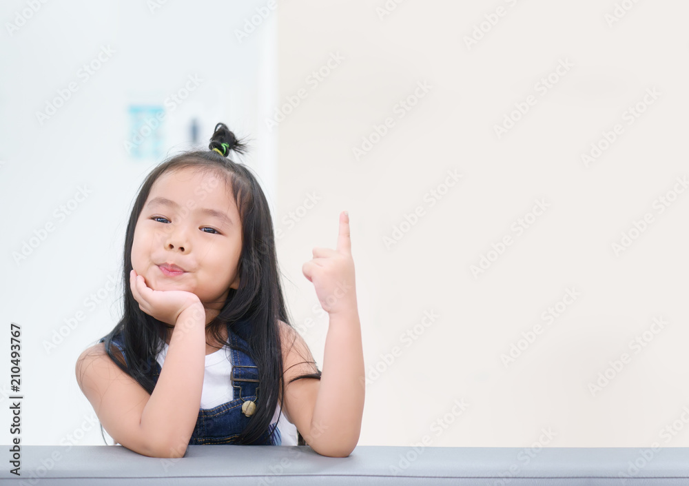 Asian children cute or kid girl thinking with finger pointing on top for new idea and happy fun or smile with wear dungarees on sofa at preschool or nursery and child hospital with space on brown Asian children cute or kid girl thinking with finger pointing on top for new idea and happy fun or smile with wear dungarees on sofa at preschool or nursery and child hospital with space on brown