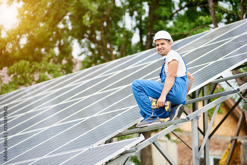 Young smiling electrician worker sitting on almost finished stand-alone solar photo voltaic panel system on bright sunny green tree background. Alternative energy concept.