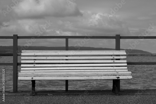 Pusta drewniana biała ławka stoi pusta na brzegu jeziora (nabrzeżu morza), za nią niska metalowa barierka, w tle woda, horyzont, chmury na niebie, czarno-białe