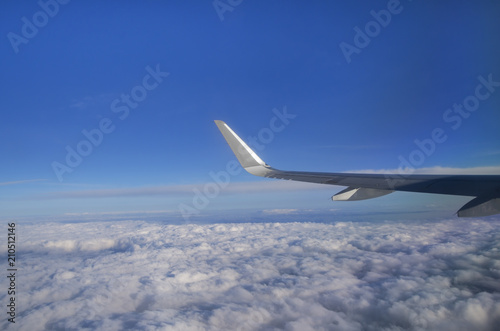 View from airplane window with silver wing, blue sky, white clouds and horizon; Flight above the clouds