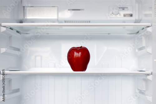 A lone red apple on a shelf in an empty fridge. One fresh apple in an empty clean refrigerator. Fruits is cooling in storage.