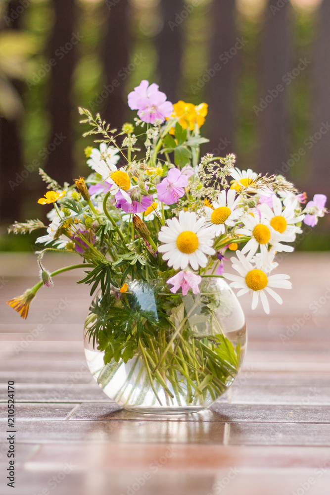 A colorful bouquet of freshly picked wild flowers from a natural meadow and garden. Presented on a table outdoors in a Clear Round Bubble Bowl Glass vase. Fence in the background.