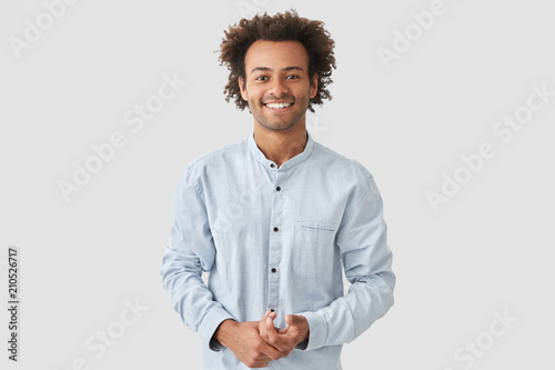 Horizontal shot of happy mixed race male with shining smile, feels glad as recieves bonus and praise for good work, has specific appearance, Afro hairstyle, dressed in fashionable white shirt