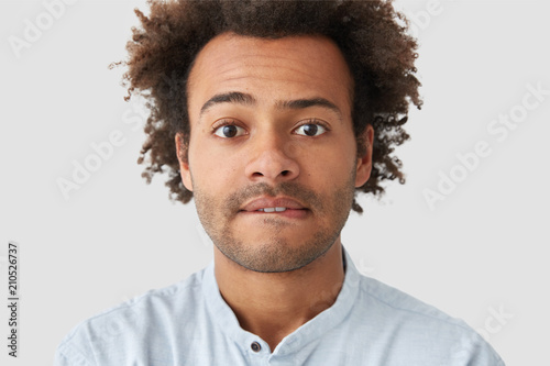 Quadro em tela Close up portrait of attractive young hipster looks embarrassed, bites lips, looks seriously at camera, has bushy hairstyle