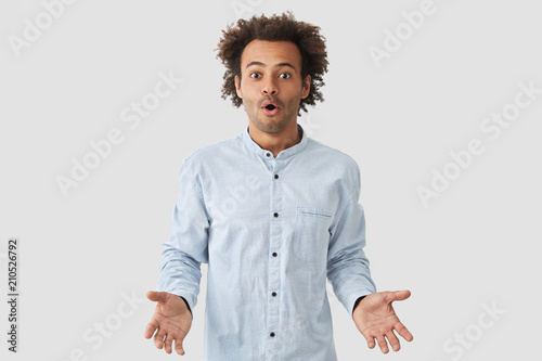 Are you kidding me? Waist up shot of attractive puzzled stupefied young African American male has dark stubble and Afro hairstyle, gestures in hesitation, dressed in white shirt, stands indoor.